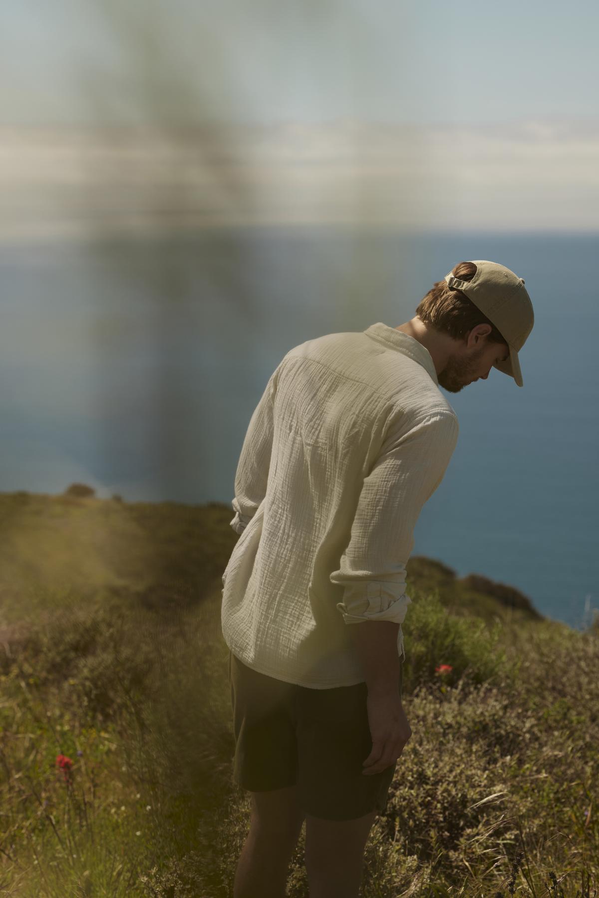   A man in a hat and light shirt looks down while standing on a coastal hill, with blurred ocean and sky in the background wearing the SALEM SHORT by Velvet by Graham & Spencer. 