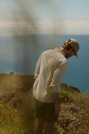 A man in a hat and light shirt looks down while standing on a coastal hill, with blurred ocean and sky in the background wearing the SALEM SHORT by Velvet by Graham & Spencer.