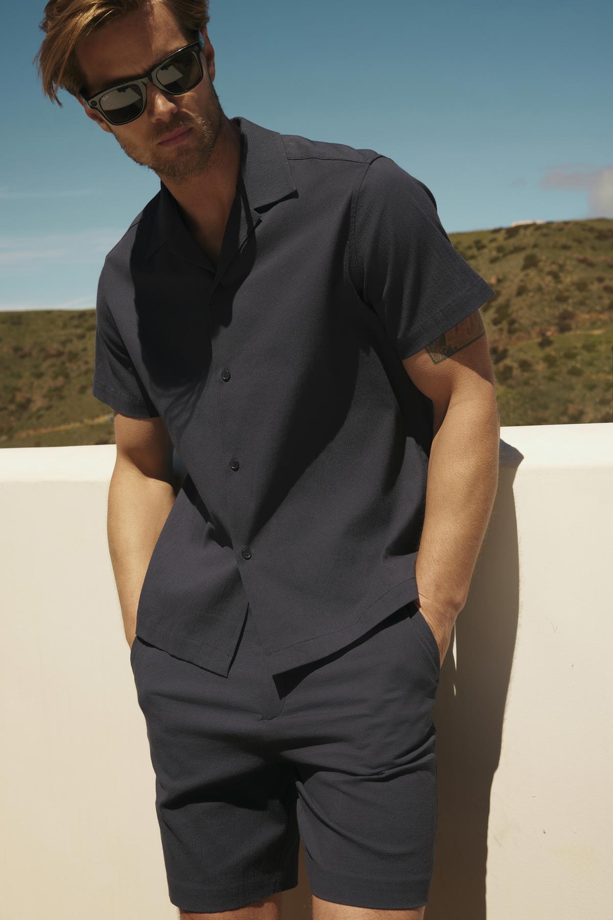   A man in sunglasses, wearing a FRANK BUTTON-UP SHIRT by Velvet by Graham & Spencer and shorts, stands against a sunlit outdoor backdrop with hills. 