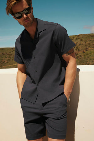 A man in sunglasses, wearing a FRANK BUTTON-UP SHIRT by Velvet by Graham & Spencer and shorts, stands against a sunlit outdoor backdrop with hills.