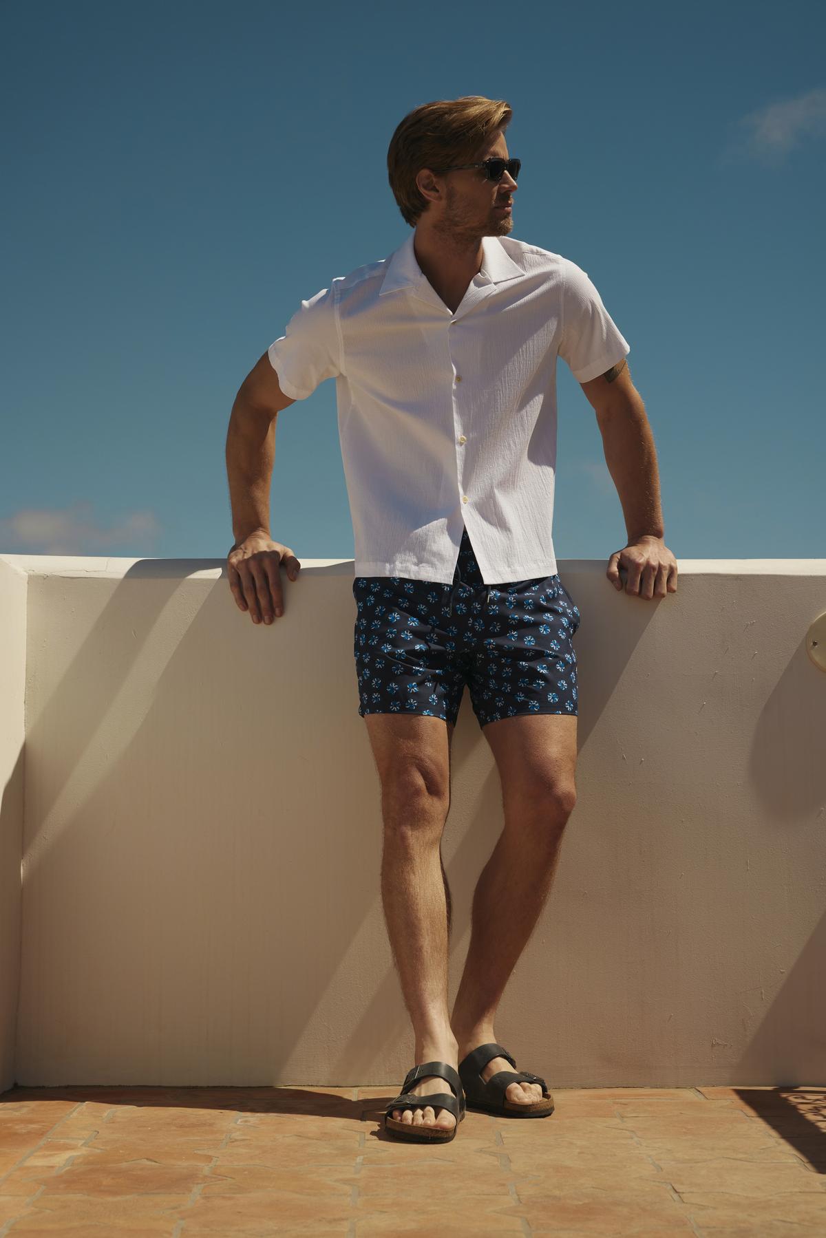 Man in sunglasses, short-sleeve white shirt, and Velvet by Graham & Spencer RICARDO SWIM SHORT standing on a sunny terrace, looking to the side.-36918671835329
