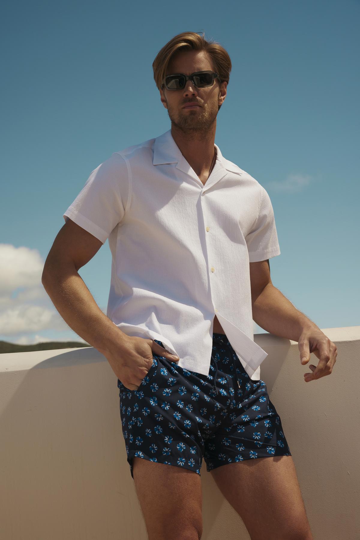   Man in sunglasses, relaxed fit white seersucker cotton FRANK BUTTON-UP SHIRT and printed shorts standing confidently against a clear sky background from Velvet by Graham & Spencer. 