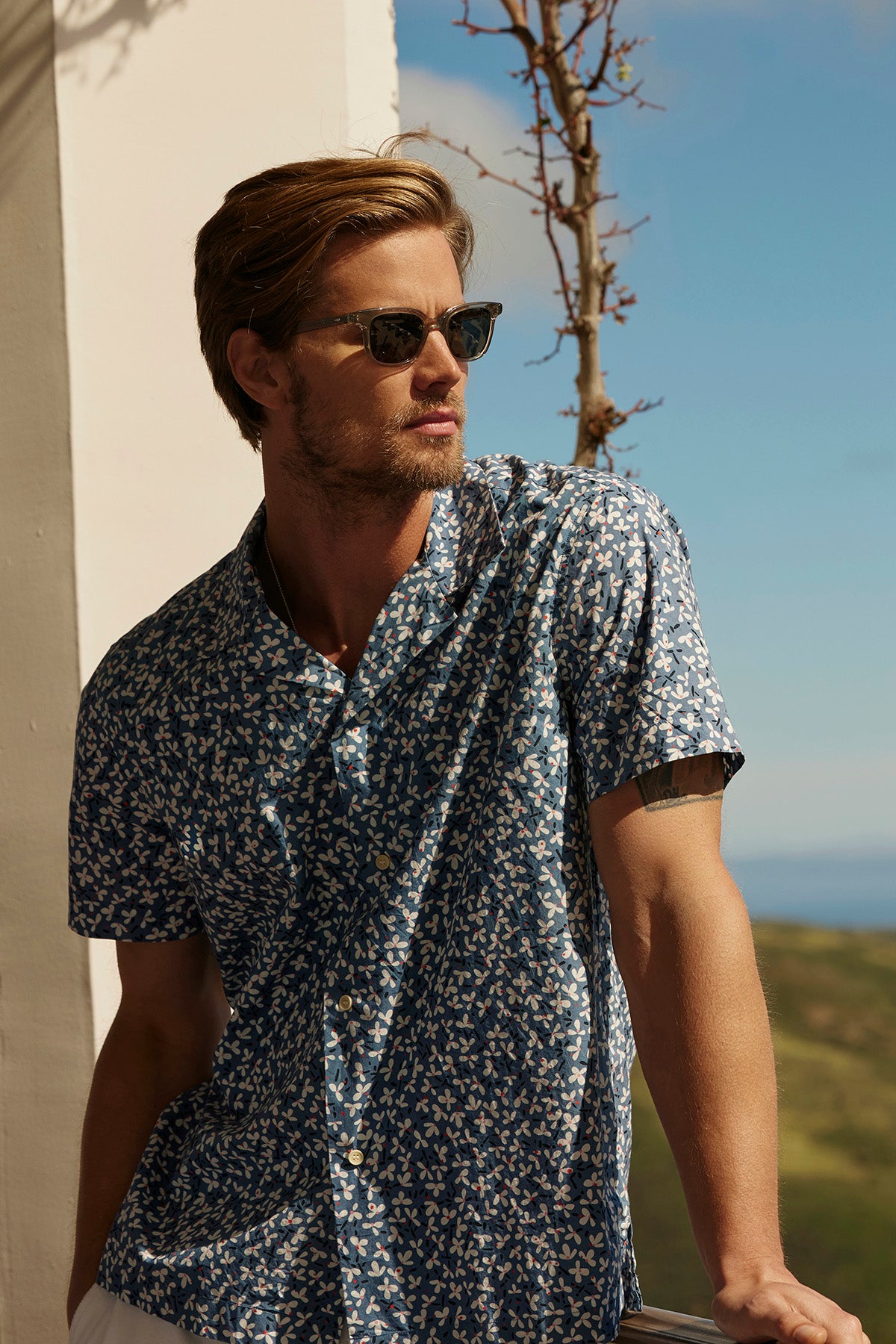   A man with light brown hair, wearing sunglasses and an IGGY BUTTON-UP SHIRT by Velvet by Graham & Spencer, featuring a blue floral design and a relaxed fit, leans against a railing with a scenic outdoor backdrop. 