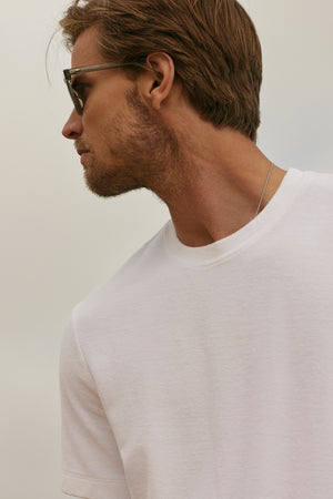 Profile view of a man with a light beard wearing sunglasses and a white cotton t-shirt with a crew neckline (JAXON CREW), looking to the left against a soft gray background.
