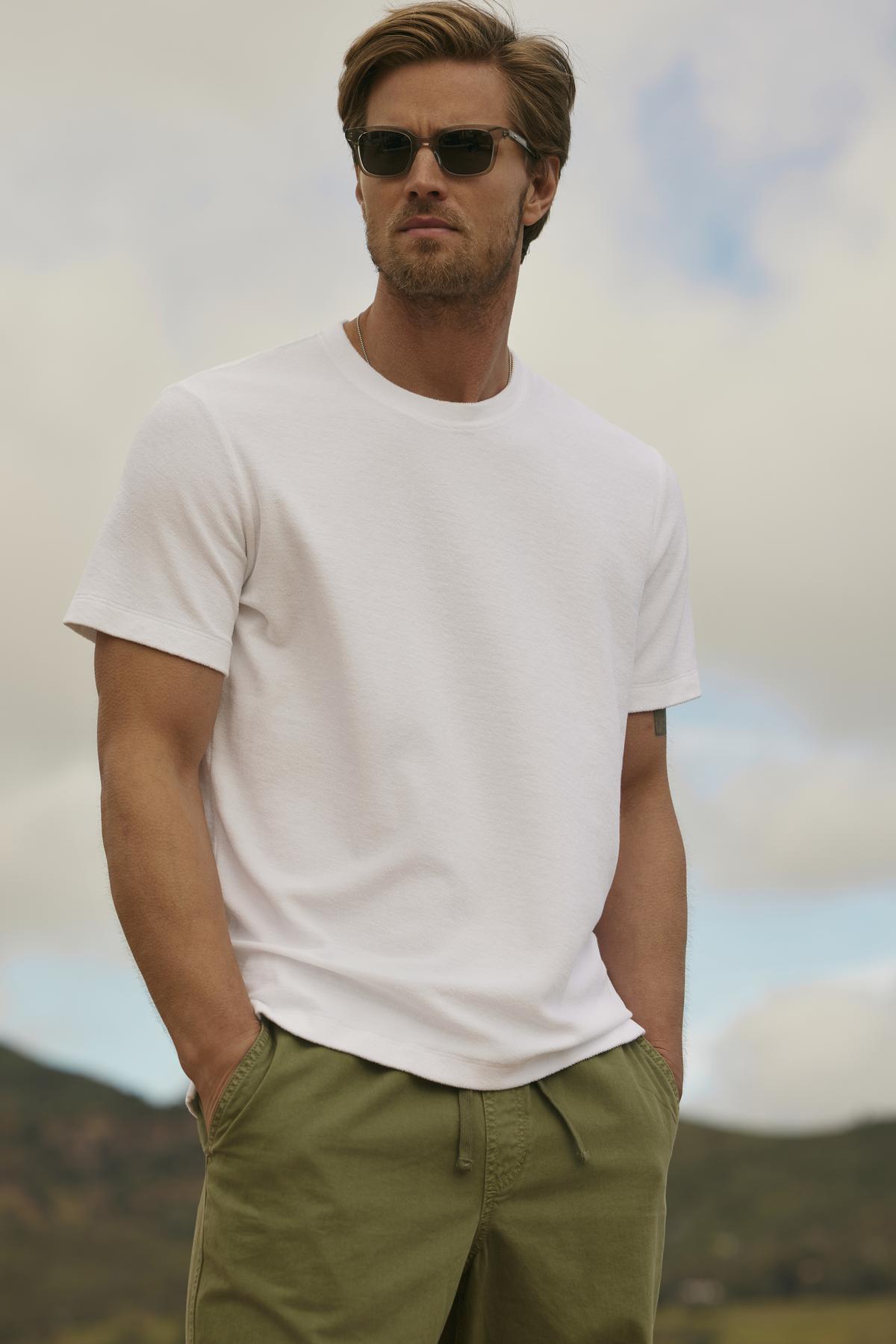 A man wearing JAXON CREW sunglasses and a white t-shirt stands with hands on hips against a hilly landscape.-36753592451265