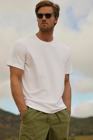 A man wearing JAXON CREW sunglasses and a white t-shirt stands with hands on hips against a hilly landscape.