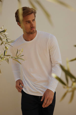 A man in a white slub knit long-sleeve shirt and black pants stands thoughtfully under soft lighting, with olive tree branches in the foreground wearing the Velvet by Graham & Spencer KAI CREW NECK TEE.