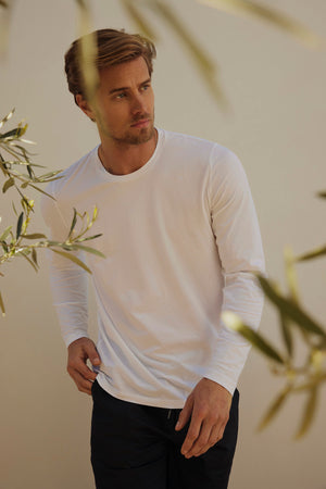 A man in a white KAI TEE by Velvet stands outdoors amidst olive branches, wearing black pants and exuding timeless design. His long-sleeve shirt features a classic crew neckline and is made from Peruvian cotton, while he gazes to the side with a neutral expression.