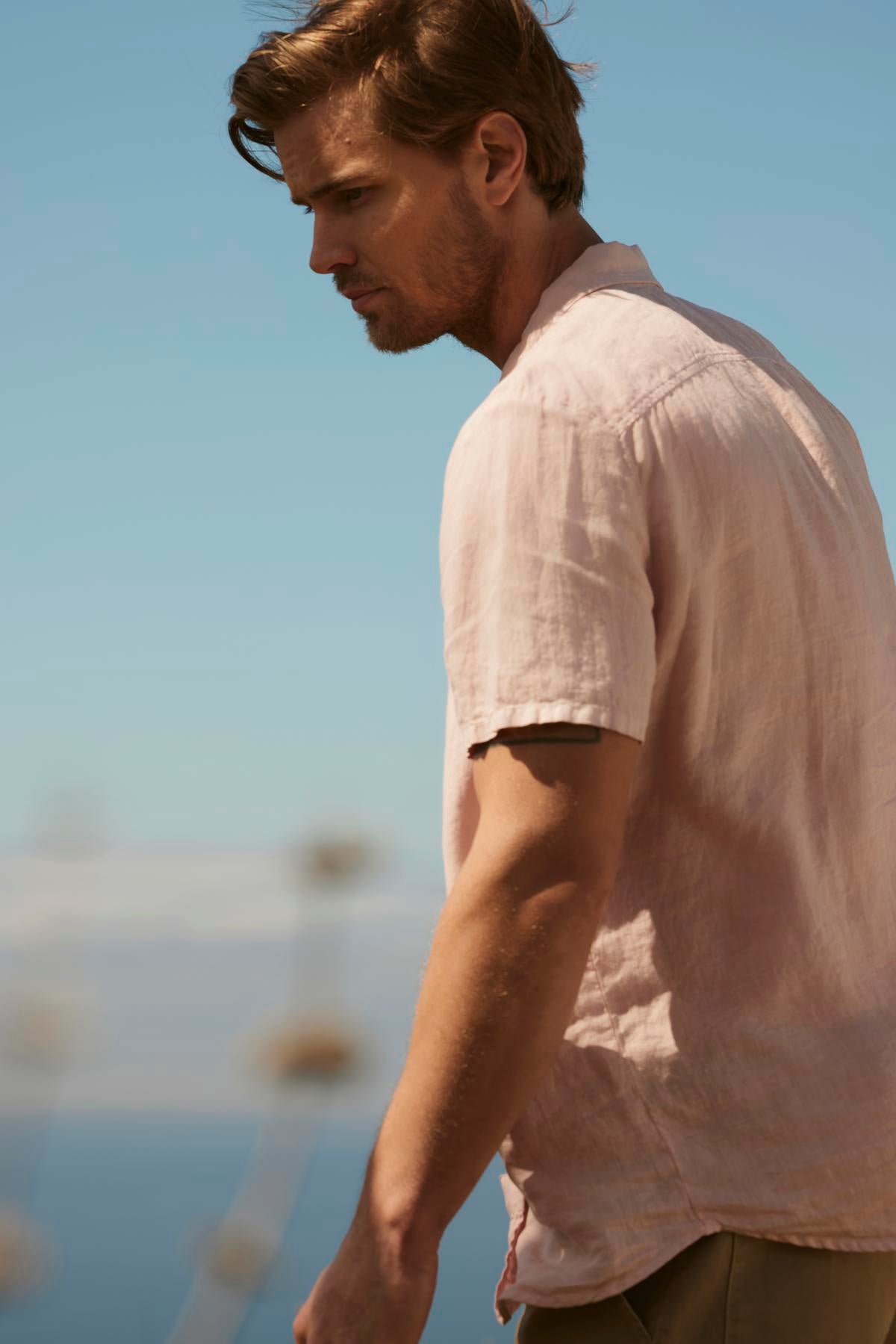 A man with light brown hair wearing a Velvet by Graham & Spencer MACKIE LINEN BUTTON-UP SHIRT gazes thoughtfully with a clear blue sky and blurry maritime background.-36753539596481