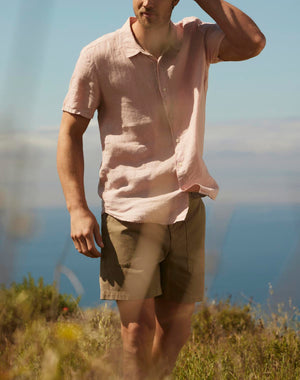 Man in a MACKIE LINEN BUTTON-UP SHIRT from Velvet by Graham & Spencer and shorts standing in a natural landscape, shielding his eyes from the sun, with a clear blue sky in the background.