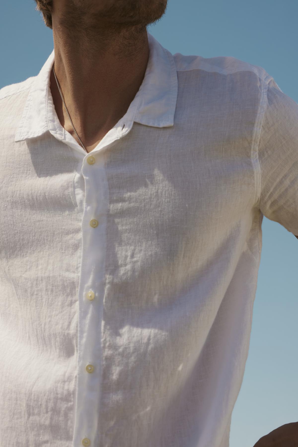   Close-up of a person wearing a Velvet by Graham & Spencer MACKIE LINEN BUTTON-UP SHIRT, focusing on the torso and unbuttoned collar. Clear blue sky in the background. 