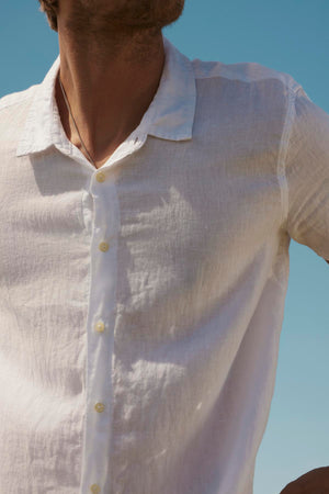 Close-up of a person wearing a Velvet by Graham & Spencer MACKIE LINEN BUTTON-UP SHIRT, focusing on the torso and unbuttoned collar. Clear blue sky in the background.
