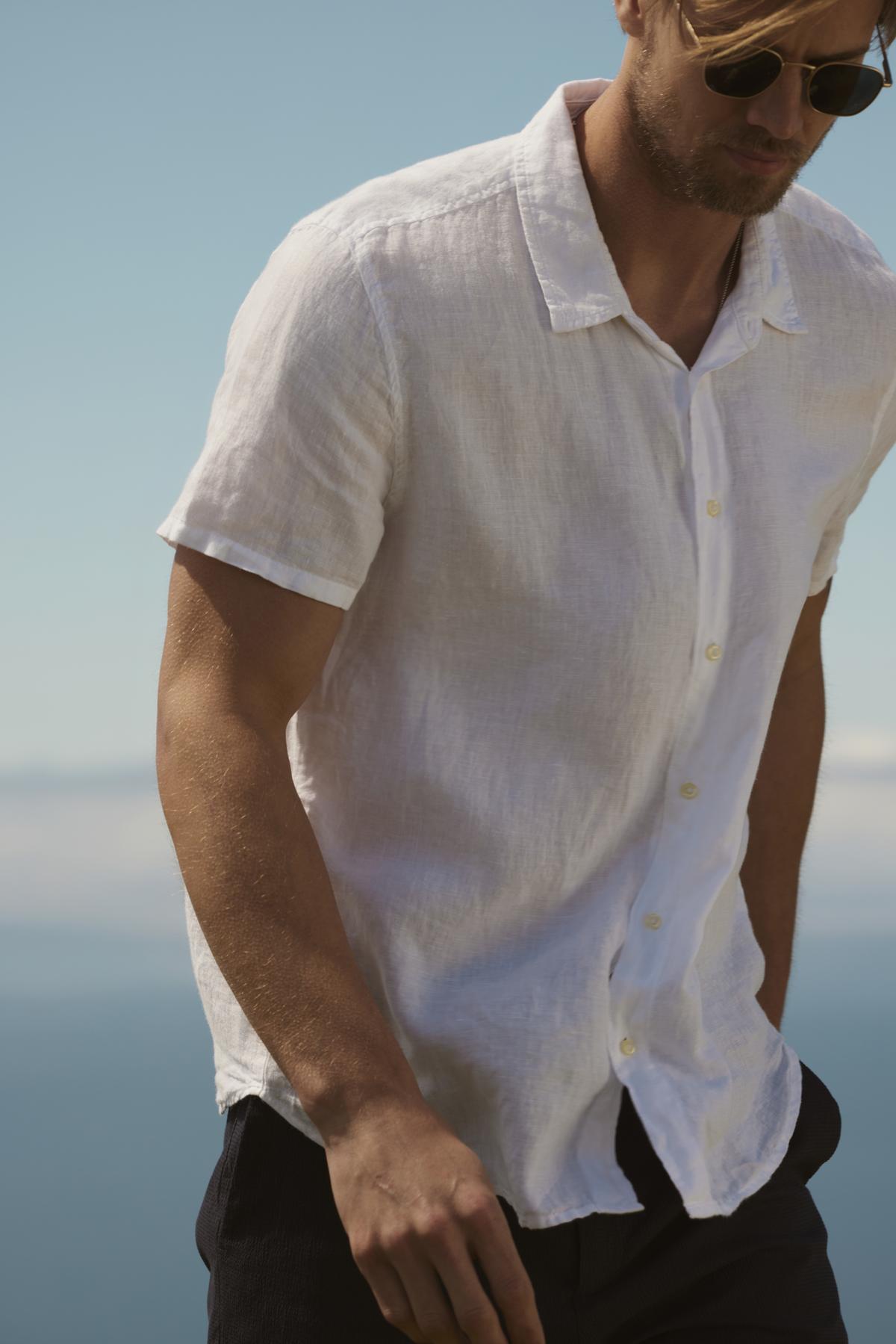A man in a white relaxed fit Velvet by Graham & Spencer MACKIE LINEN BUTTON-UP SHIRT and sunglasses stands by the sea, a clear sky in the background.-36753542643905