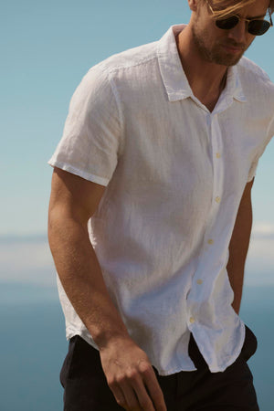 A man in a white relaxed fit Velvet by Graham & Spencer MACKIE LINEN BUTTON-UP SHIRT and sunglasses stands by the sea, a clear sky in the background.