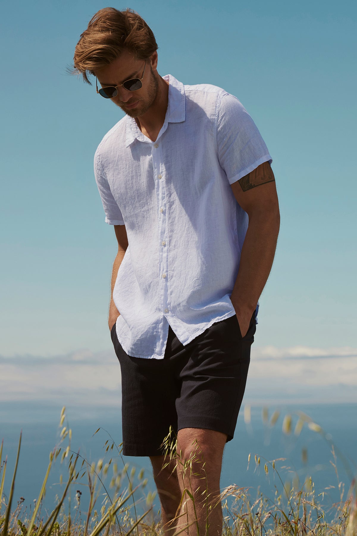 Man in a Velvet by Graham & Spencer MACKIE LINEN BUTTON-UP SHIRT and black shorts standing in a grassy area with the ocean in the background on a sunny day.-36909328564417