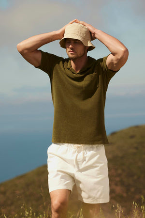 A man wearing an olive green shirt, beige bucket hat, and Velvet by Graham & Spencer SALEM SHORT with a drawstring elastic waist is standing outdoors with a hand on his head. The background includes hills and a cloudy sky, making it perfect for casual outings.