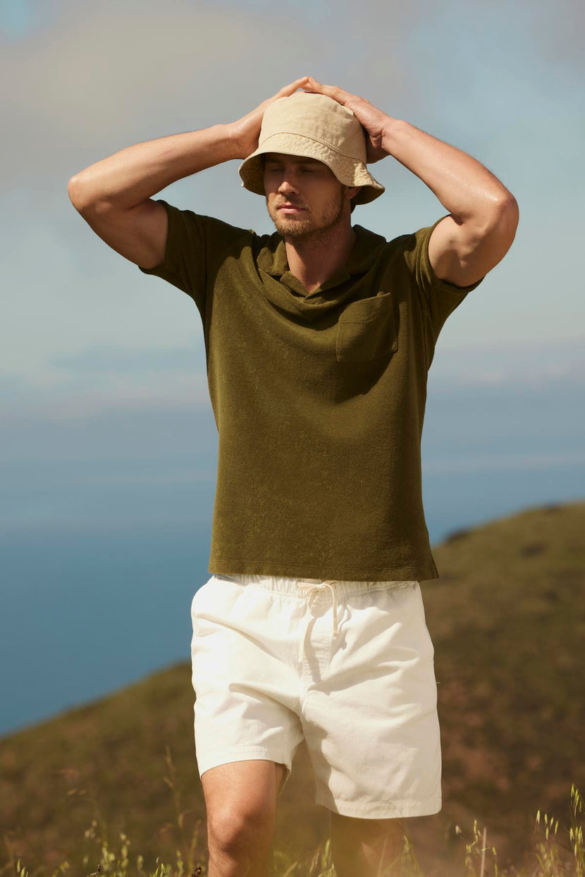 A man wearing FIELDER SHORT by Velvet by Graham & Spencer holds a wide-brimmed hat on his head while standing in a green field with a clear blue sky and distant hills.