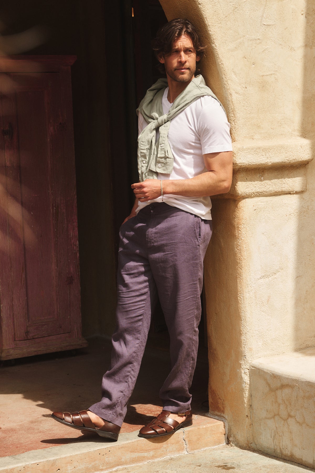 A man stands against a stucco wall wearing Velvet by Graham & Spencer’s ALFRED LINEN PANT—straight-leg, breathable linen trousers with a rear elastic waistband—paired with a white T-shirt, light sweater over his shoulders, and brown sandals.-39152330440897