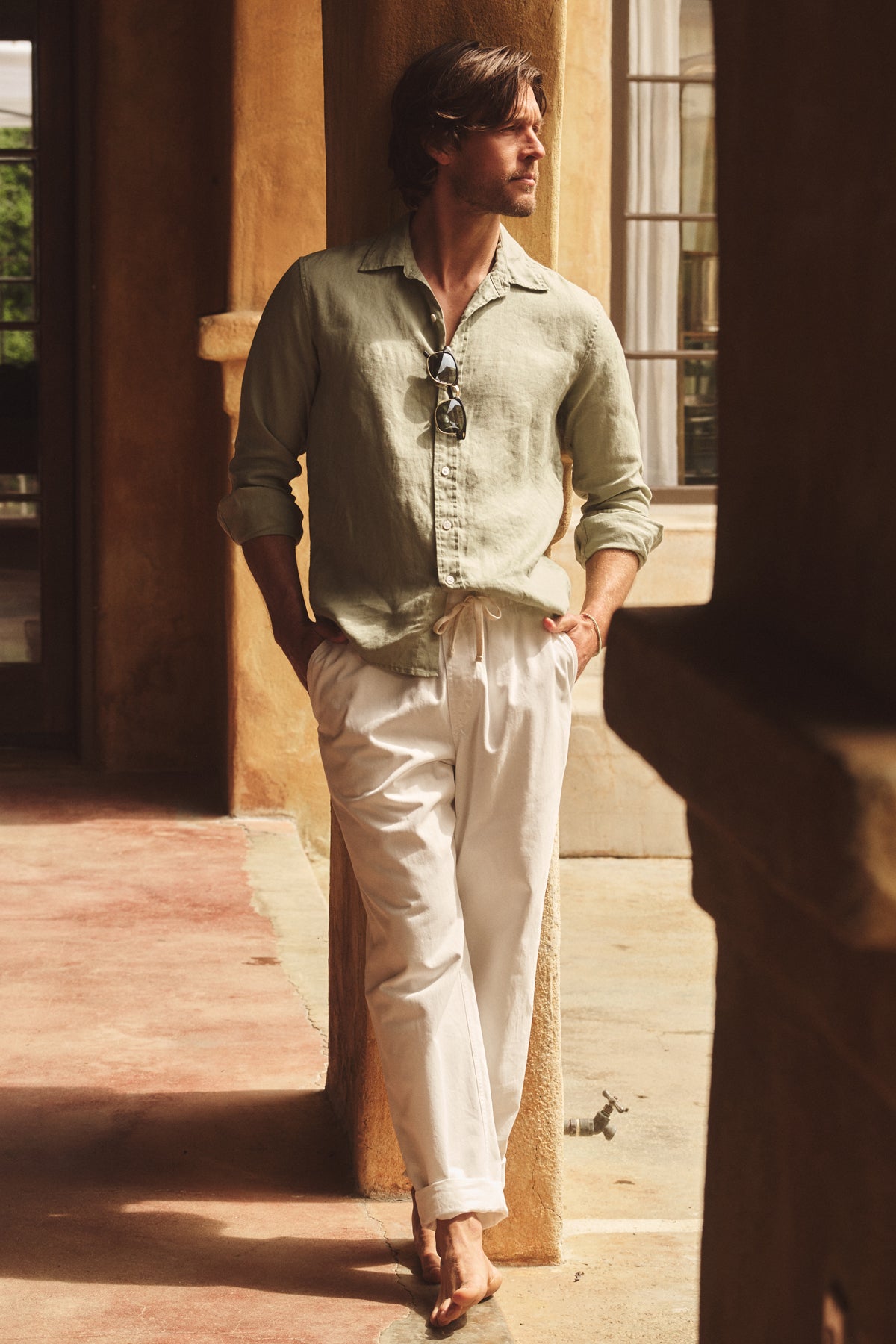 A man stands barefoot in a shaded outdoor corridor, hands in pockets, wearing the Velvet by Graham & Spencer CARUSO LINEN BUTTON-UP SHIRT and white pants—sunglasses hanging from his shirt highlight this classic summer look.-39152342466753