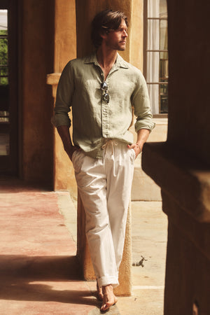 A man stands barefoot in a shaded outdoor corridor, hands in pockets, wearing the Velvet by Graham & Spencer CARUSO LINEN BUTTON-UP SHIRT and white pants—sunglasses hanging from his shirt highlight this classic summer look.