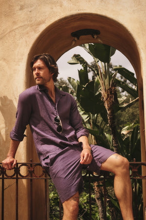 A man wearing the CARUSO LINEN BUTTON-UP SHIRT by Velvet by Graham & Spencer and shorts sits on a railing under an archway, with sunglasses hanging from his shirt and lush tropical plants in the background.