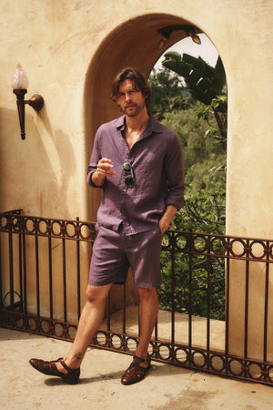 A man stands by an arched doorway wearing the Velvet by Graham & Spencer CARUSO LINEN BUTTON-UP SHIRT, one hand in his pocket and sunglasses in the other, highlighting this versatile style.