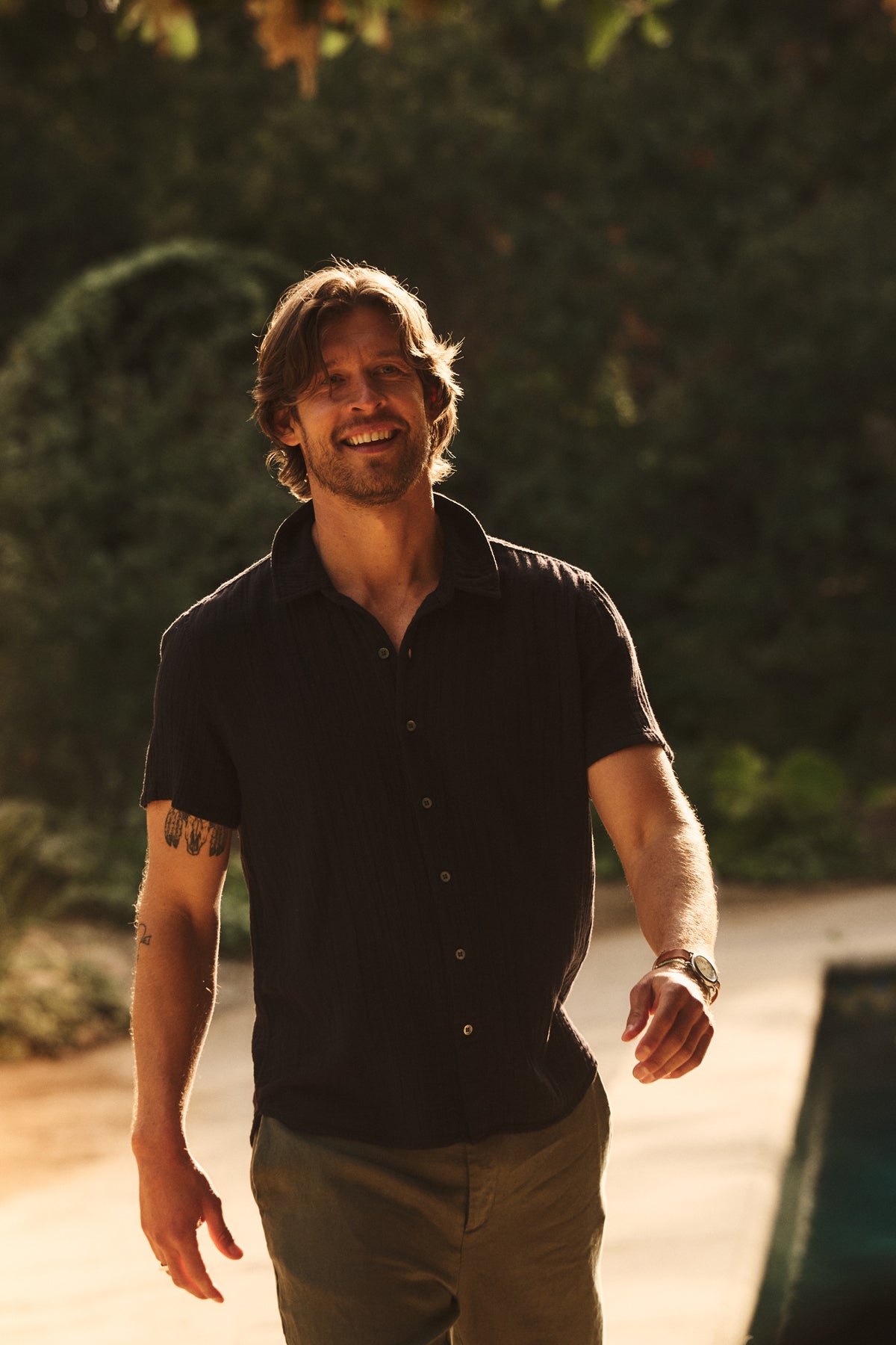   A man with mid-length hair and a beard smiles as he walks outdoors near greenery, wearing the CHRISTIAN COTTON GAUZE BUTTON-UP SHIRT by Velvet by Graham & Spencer with pants. 