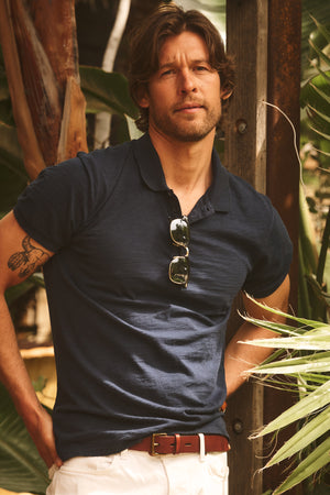 A man with wavy hair, dressed in the Velvet by Graham & Spencer NIKO POLO and white pants, sunglasses hanging from his collar, stands outdoors near plants and looks at the camera.
