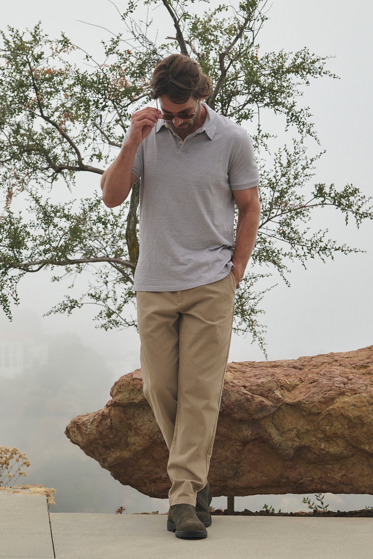   A man wearing the MORAN PANT from Velvet by Graham & Spencer, paired with a gray polo, strolls outdoors while adjusting his sunglasses. In the background, a large rock and tree can be seen. 