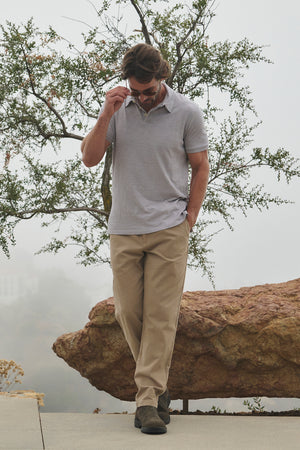 A man wearing the MORAN PANT from Velvet by Graham & Spencer, paired with a gray polo, strolls outdoors while adjusting his sunglasses. In the background, a large rock and tree can be seen.