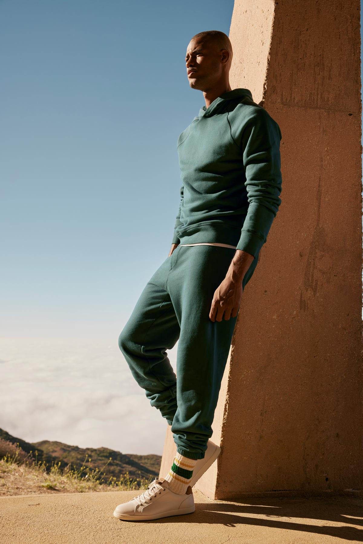   A man in a green hoodie and MONTGOMERY BRUSHED FLEECE SWEATPANTS by Velvet by Graham & Spencer leaning against a wall. 