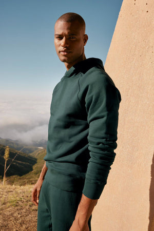 A man in a Green Velvet by Graham & Spencer Hopkin Pullover Hoodie experiencing superior comfort on top of a mountain.