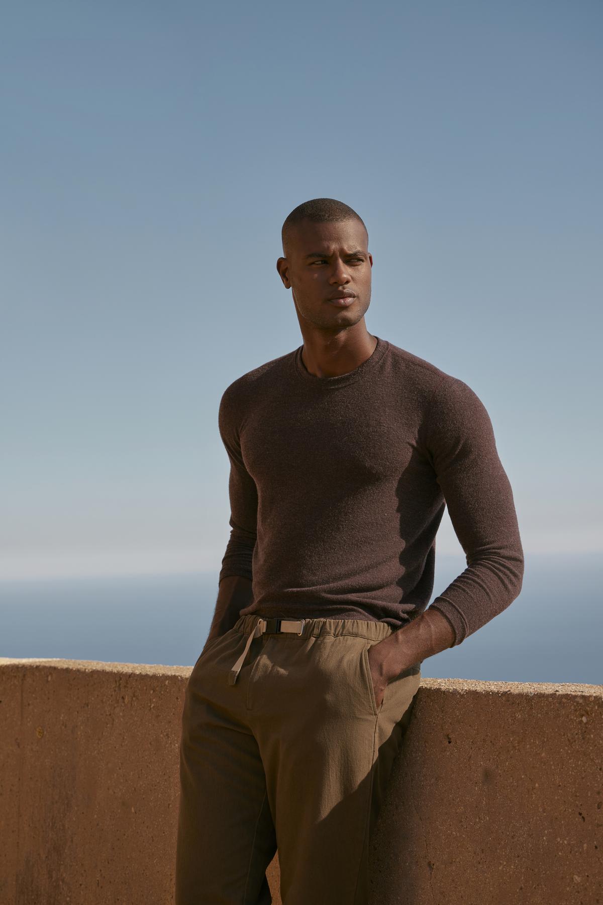 A man in a Velvet by Graham & Spencer cozy brown sweater, the BECKER COZY JERSEY CREW, and soft khaki pants leaning against a wall.-35548082897089