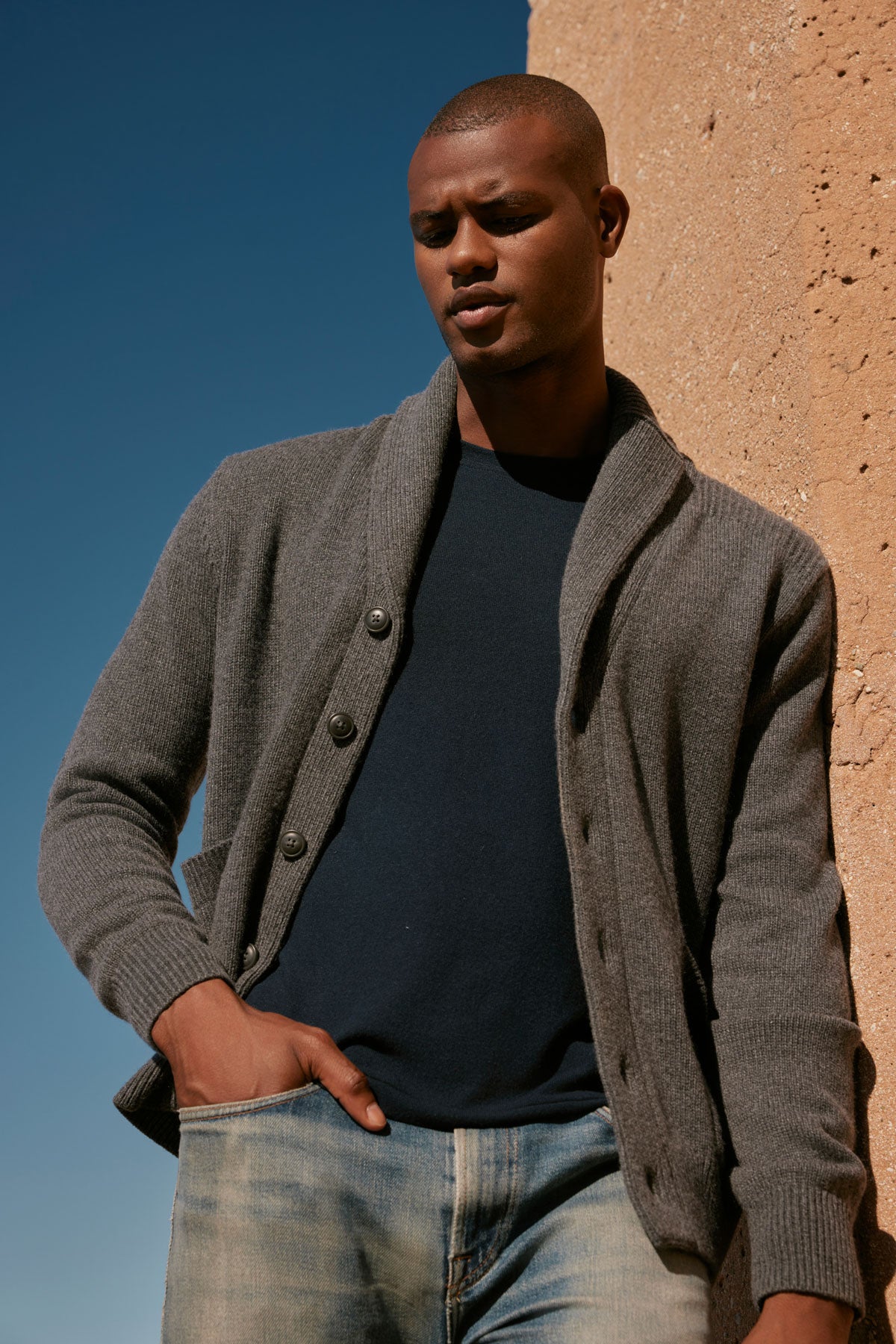 A man in jeans and a polished appearance is leaning against a wall wearing the Velvet by Graham & Spencer THORNTON BUTTON FRONT CARDIGAN.-35605827223745