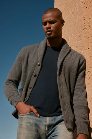 A man in jeans and a polished appearance is leaning against a wall wearing the Velvet by Graham & Spencer THORNTON BUTTON FRONT CARDIGAN.