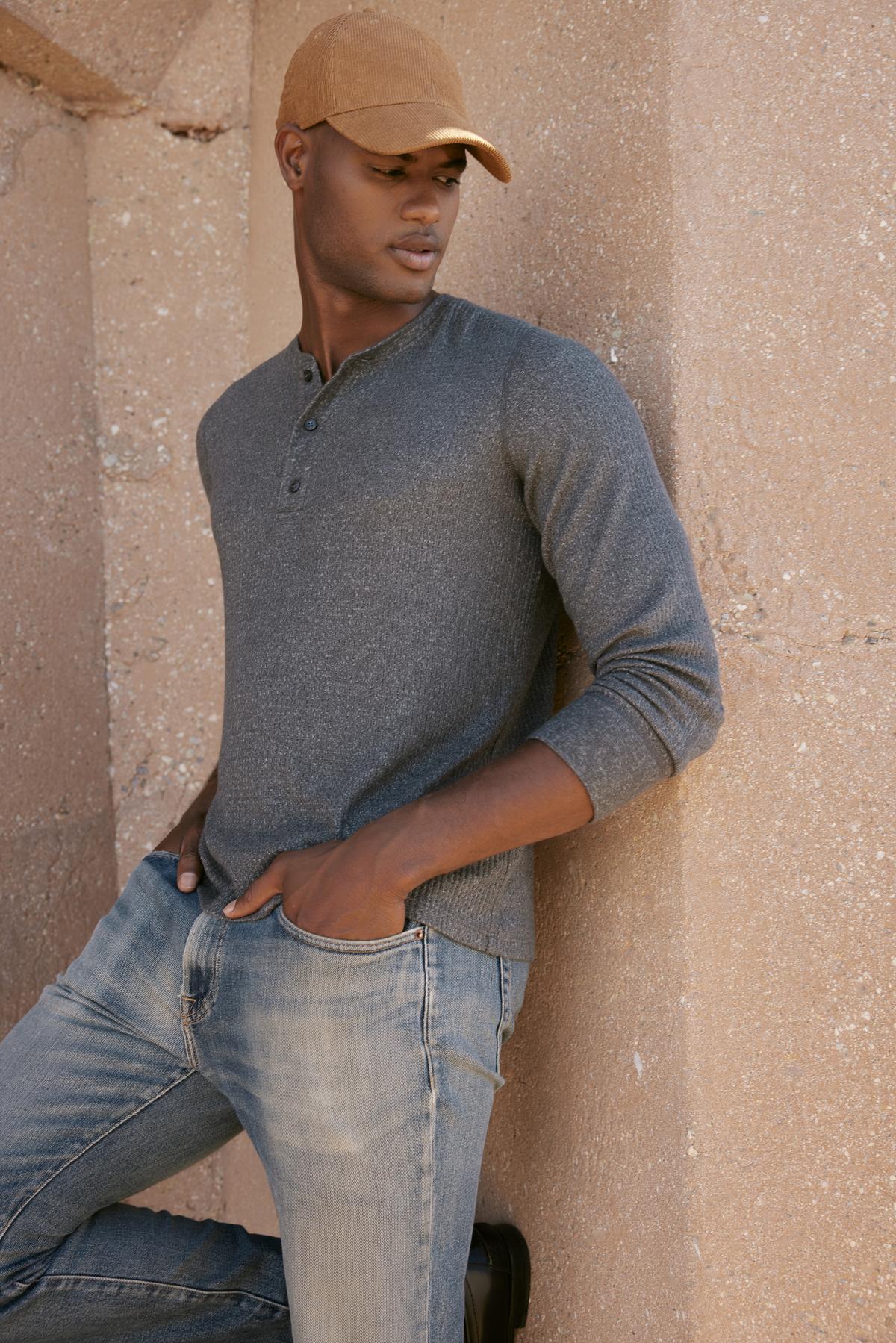   A man in jeans and a hat leaning against a wall, showcasing the Velvet by Graham & Spencer's ANTHONY THERMAL HENLEY's classic waffle texture. 