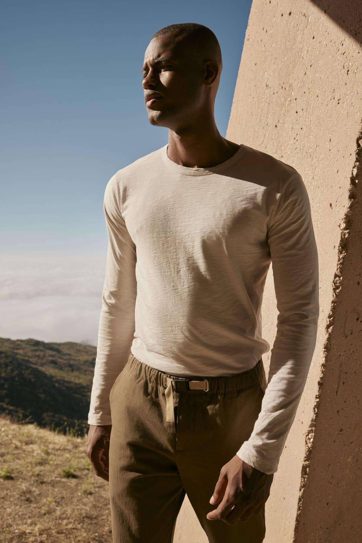   A man in a white KAI CREW NECK TEE by Velvet by Graham & Spencer and khaki pants is standing in front of a mountain. 