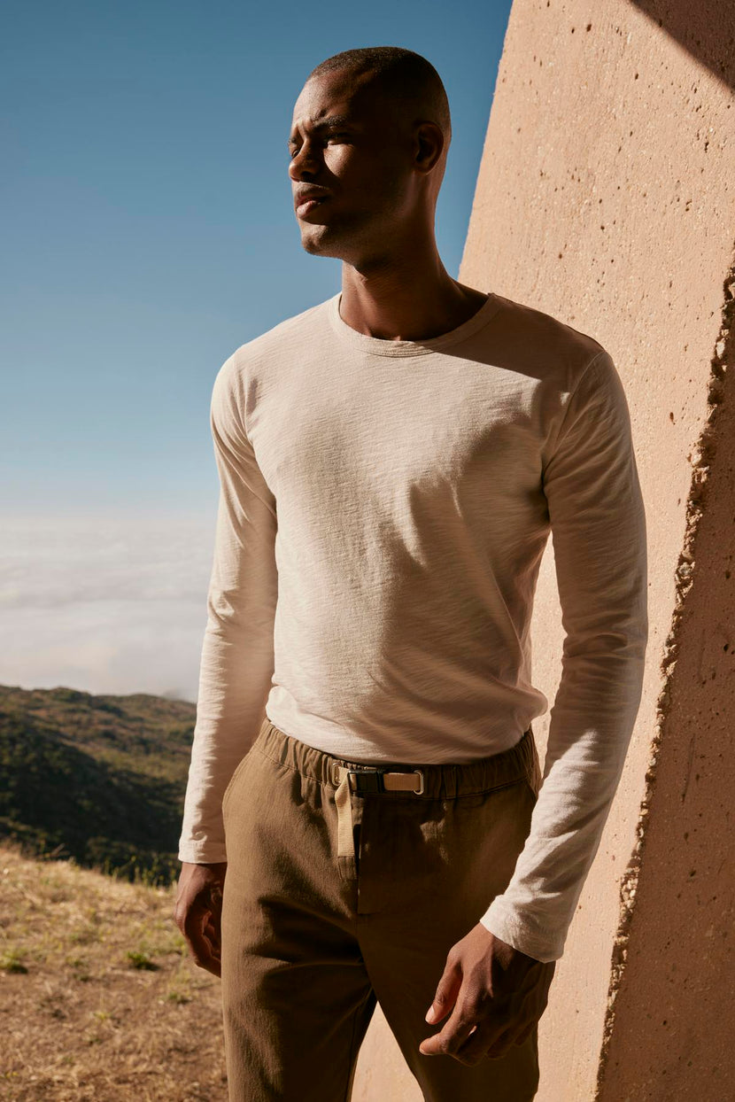 A man in a white KAI CREW NECK TEE by Velvet by Graham & Spencer and khaki pants is standing in front of a mountain.