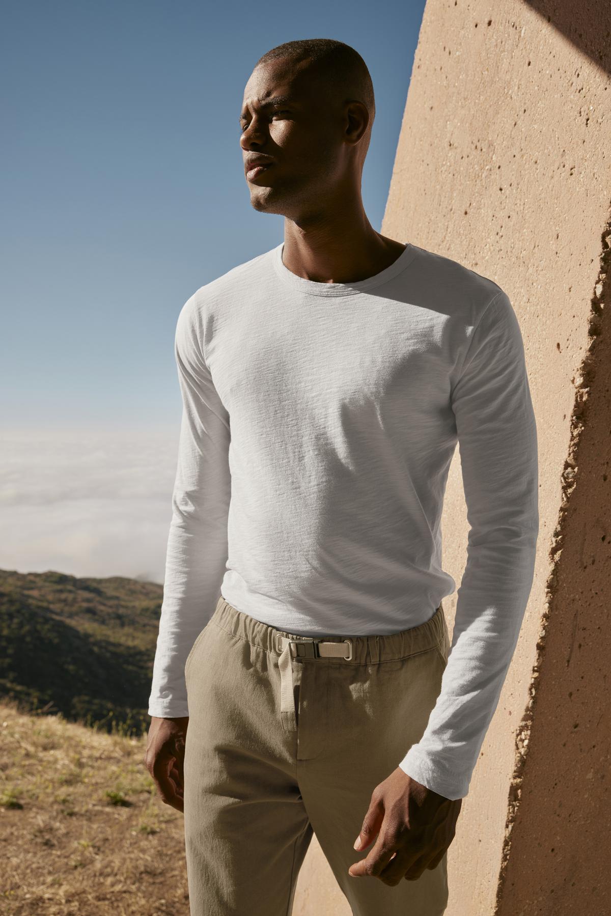   A person stands outdoors wearing a long-sleeve white shirt and Velvet by Graham & Spencer's MORAN PANT, set against the backdrop of a concrete structure and a scenic landscape. 