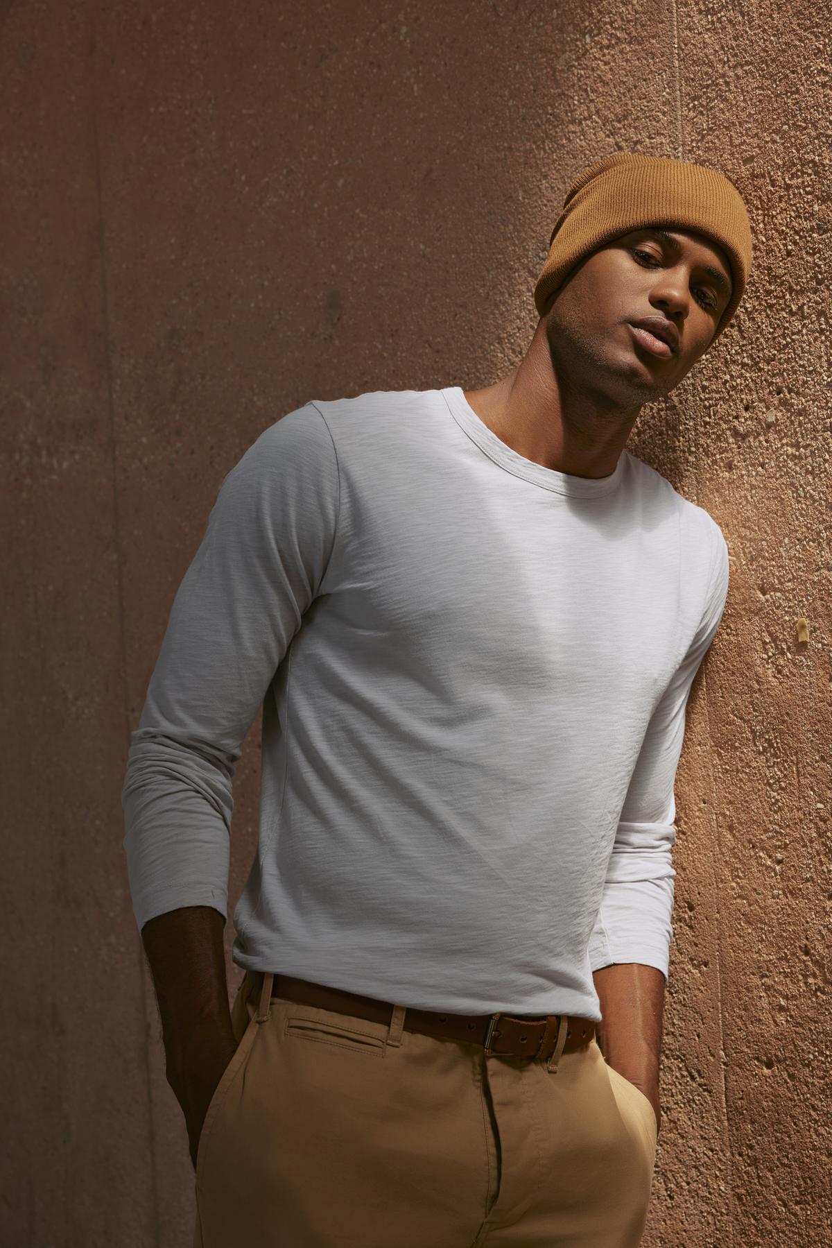 A man in the KAI TEE by Velvet by Graham & Spencer, a white long-sleeve shirt crafted from lightweight Peruvian cotton, and a brown beanie leans against a textured wall, showcasing timeless design.-38264831279297