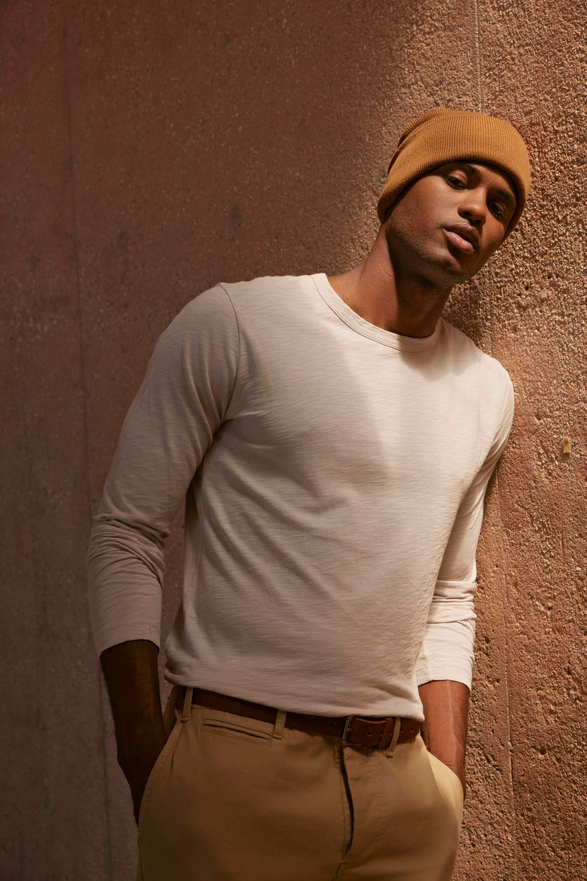 A man wearing a beanie, leaning against a wall, with a vintage-feel texture was wearing the KAI CREW NECK TEE by Velvet by Graham & Spencer.