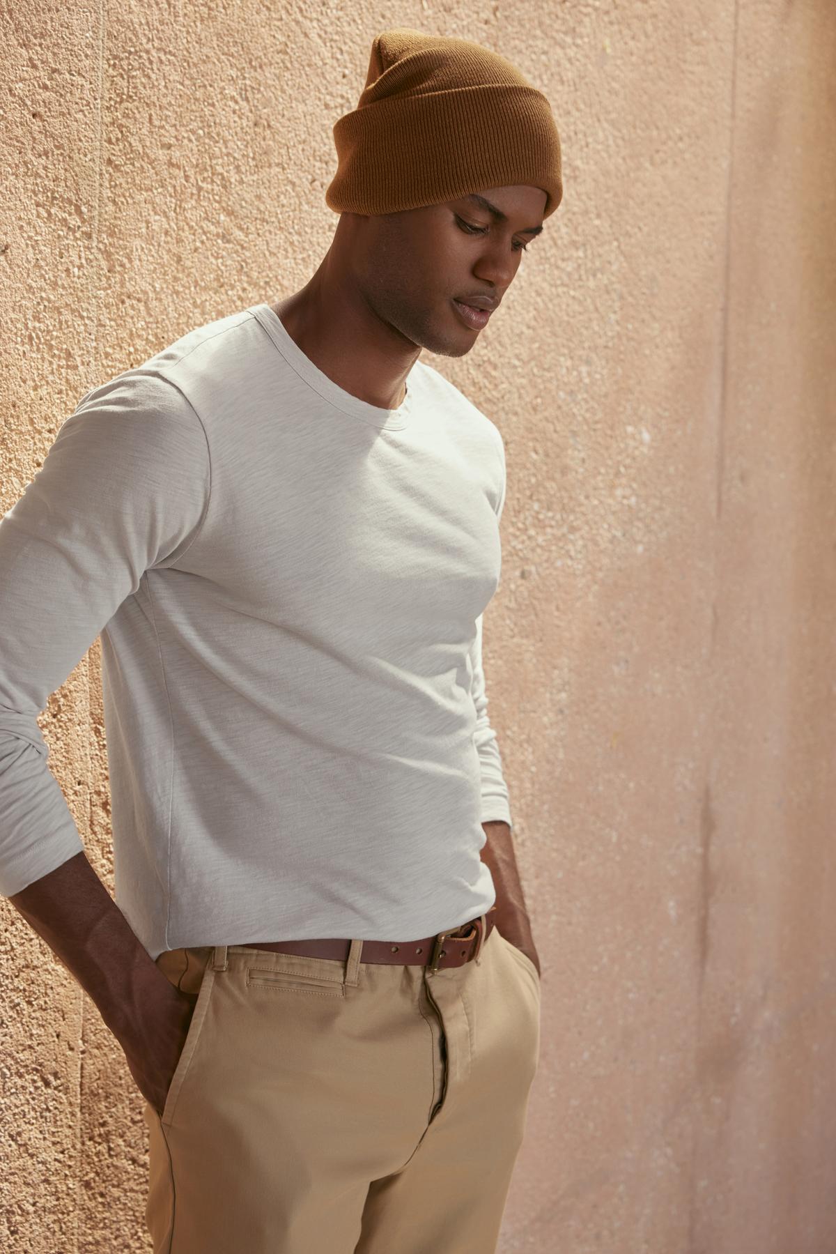 A person dressed in the KAI TEE by Velvet by Graham & Spencer, along with beige pants and a brown beanie, stands against a textured wall, highlighting a timeless design.-38264831967425