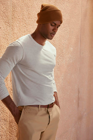 A person dressed in the KAI TEE by Velvet by Graham & Spencer, along with beige pants and a brown beanie, stands against a textured wall, highlighting a timeless design.