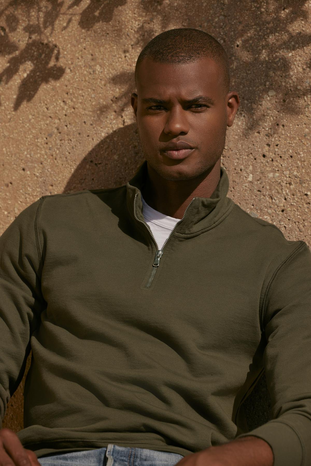 A man in a green Velvet by Graham & Spencer BANNING QUARTER-ZIP SWEATSHIRT leaning against a wall.-35547504509121