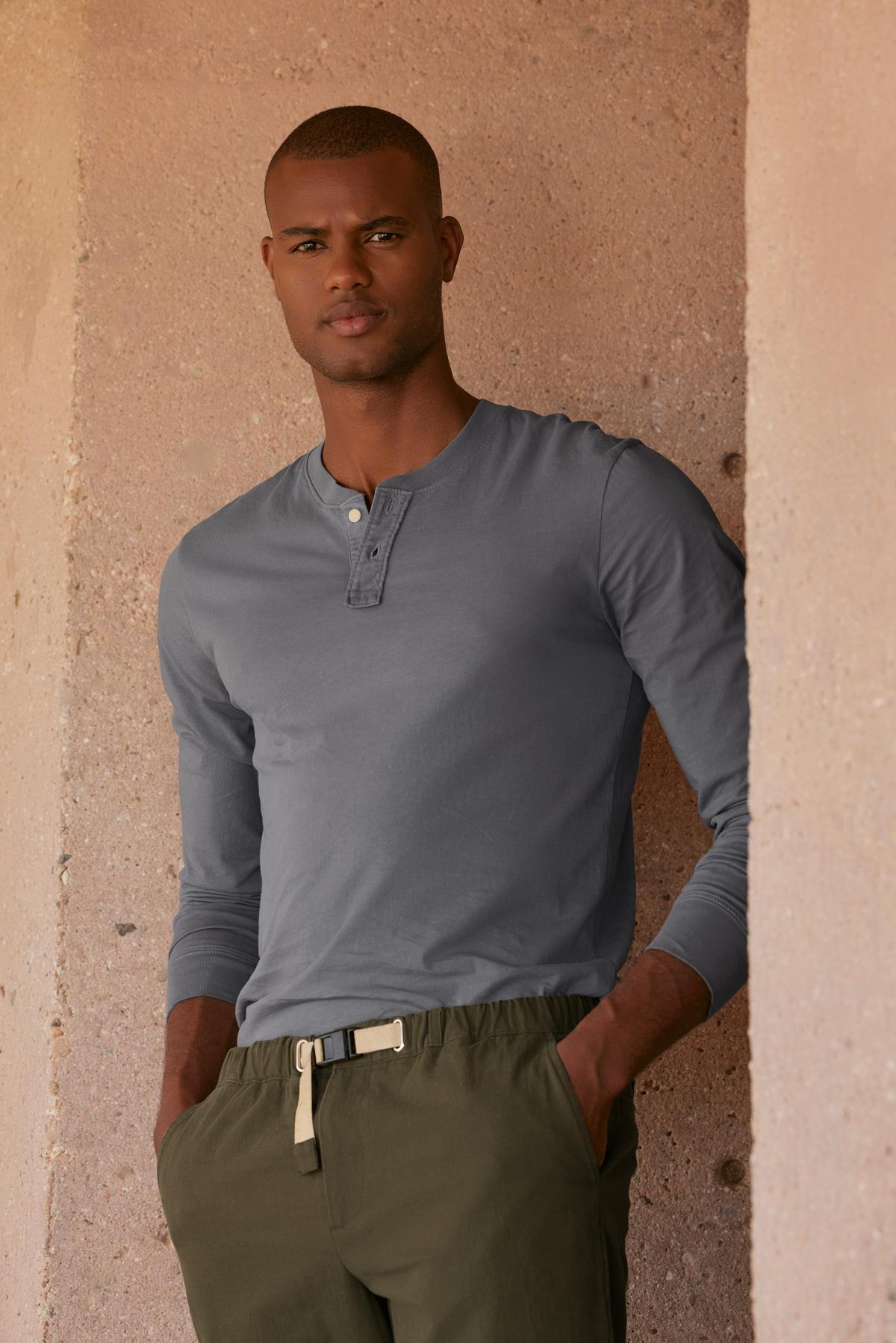   A man wearing a gray BRADEN HENLEY shirt by Velvet by Graham & Spencer is casually leaning against a wall. 