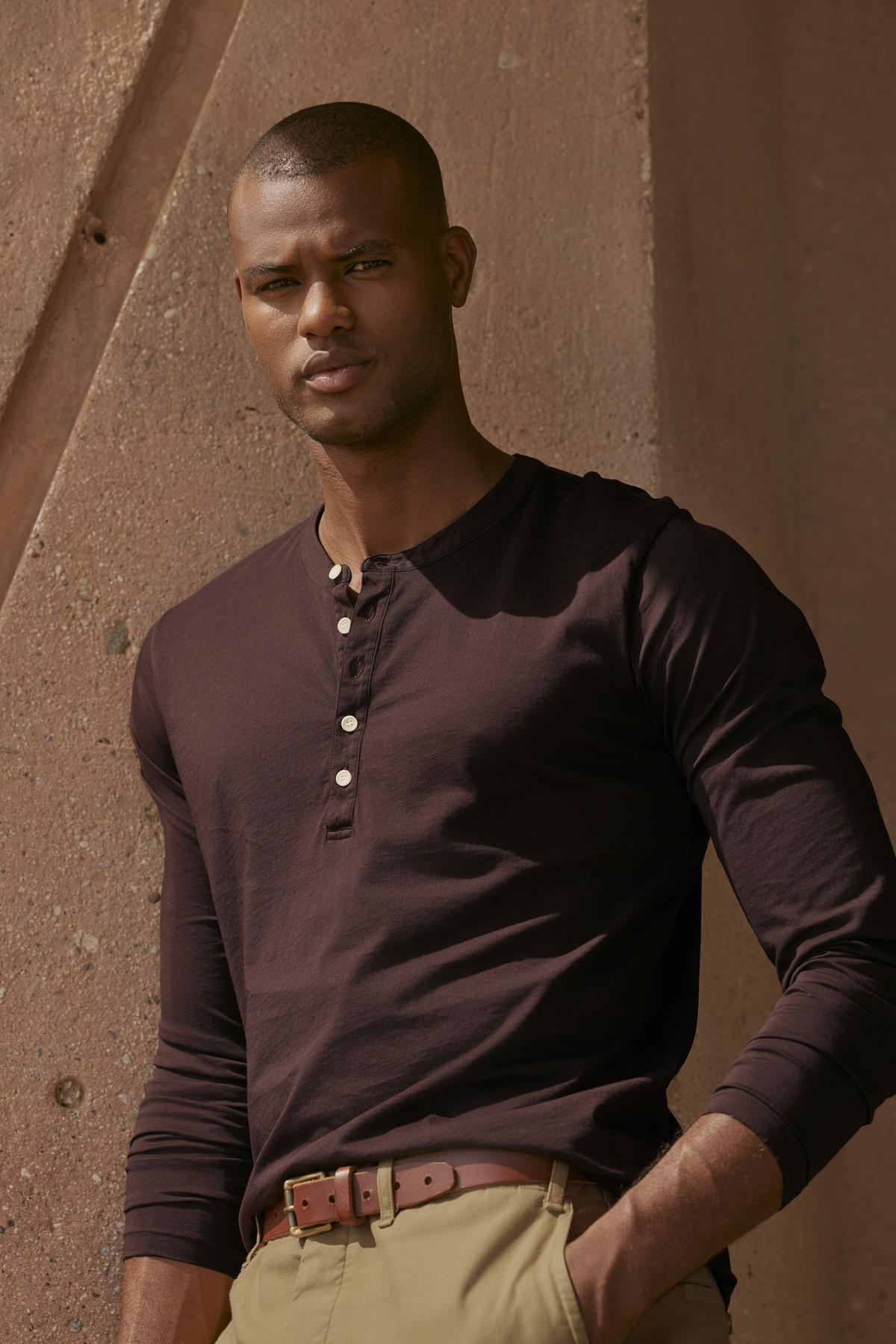 A man in a brown shirt and khaki pants is leaning against a wall, wearing a vintage-look Velvet by Graham & Spencer ALVARO COTTON JERSEY HENLEY.-35547510309057