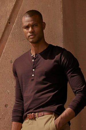 A man in a brown shirt and khaki pants is leaning against a wall, wearing a vintage-look Velvet by Graham & Spencer ALVARO COTTON JERSEY HENLEY.