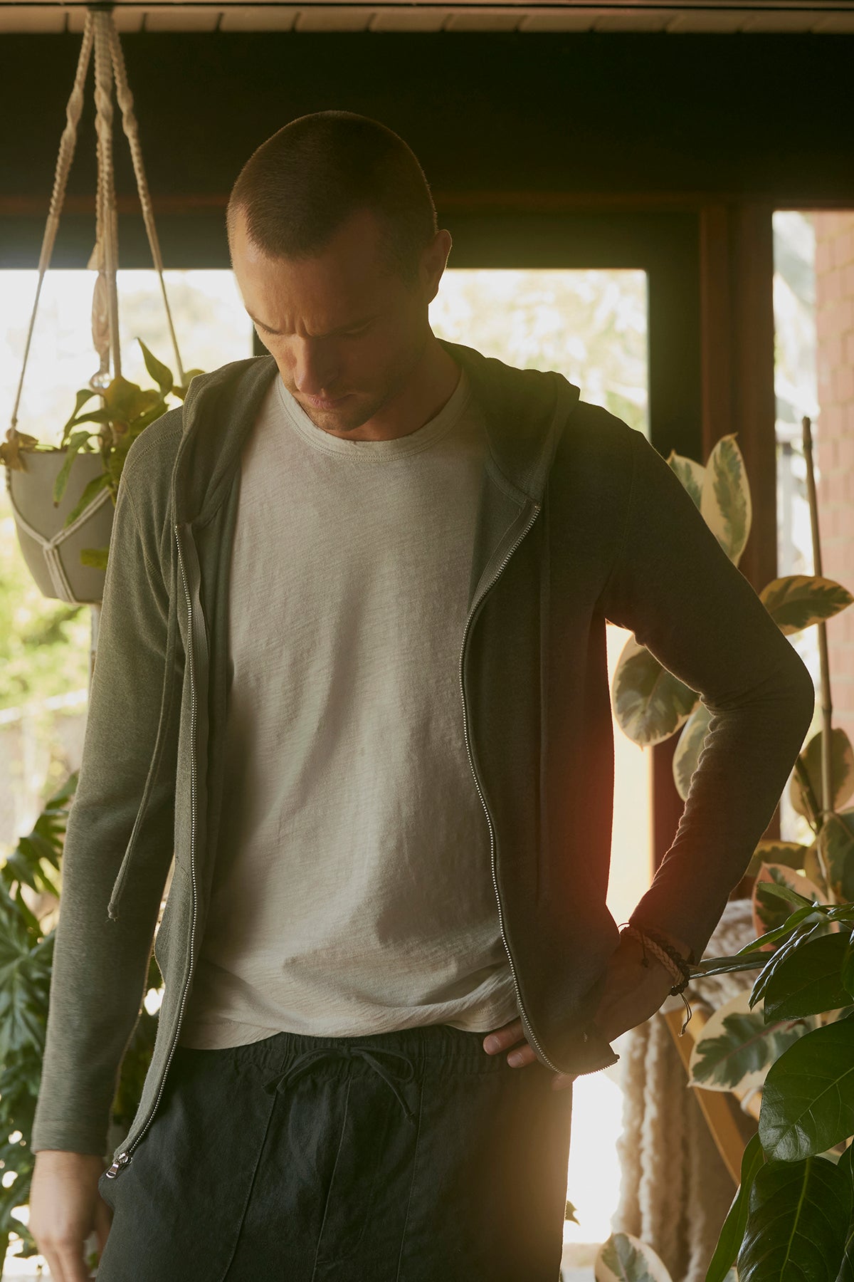 A man in casual clothing from Velvet by Graham & Spencer stands indoors near houseplants with a neutral expression, wearing the AMARO TEE. The classic crew neckline and lightweight Peruvian cotton of his shirt add a touch of comfort to his relaxed look.-37759204720833