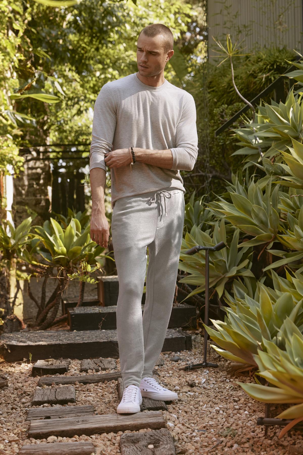   A man in a light gray long-sleeve shirt and Velvet by Graham & Spencer's CROSBY JOGGERs stands on a stone path, surrounded by greenery and wooden steps, looking off to the side. 