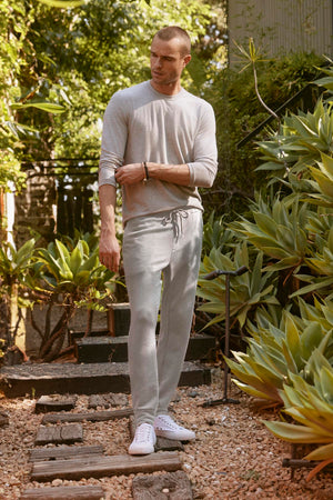 A man in a light gray long-sleeve shirt and Velvet by Graham & Spencer's CROSBY JOGGERs stands on a stone path, surrounded by greenery and wooden steps, looking off to the side.